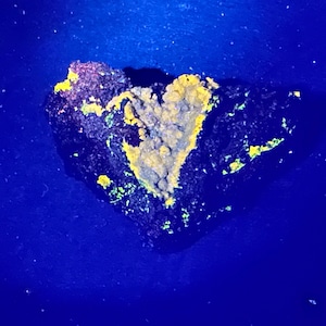 May include: A heart-shaped rock formation with a dark blue base, speckled with small white particles. The rock has a rough texture, with yellow and green mineral deposits. The top of the rock has a pinkish hue.