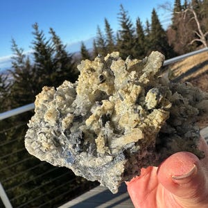 Może przedstawiać: Dłoń trzyma skupisko surowych minerałów. Formacja mineralna charakteryzuje się mieszanką kolorów, w tym beżowym, szarym i ciemnoniebieskim. Tło przedstawia błękitne niebo i wiecznie zielone drzewa.