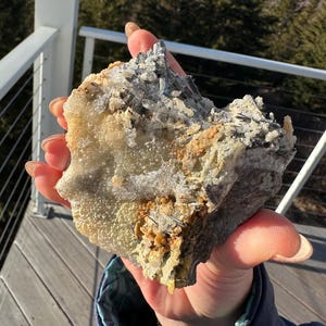 Może przedstawiać: Dłoń trzyma szorstki, naturalny okaz minerału. Skała ma mieszankę kolorów, w tym beżowy, szary i odcienie pomarańczowego. Minerał ma krystaliczną strukturę z widocznymi formacjami. Tło przedstawia drewniany taras i widok na drzewa.