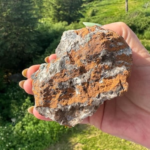 Op de afbeelding: Een ruwe, onregelmatig gevormde steen met lagen van oranje, grijze en witte mineralen. De steen wordt in een hand gehouden, met een groen gelakte nagel zichtbaar. De achtergrond toont groene bomen en gras.