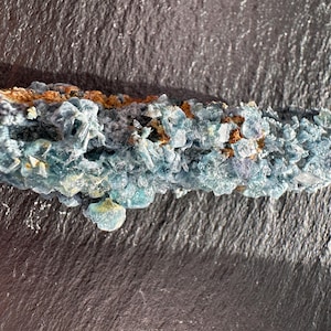 May include: A close-up of a mineral specimen featuring a cluster of blue-green crystals with hints of purple and brown. The crystals have a rough, textured surface and are arranged in a compact formation. The mineral is set against a dark background.