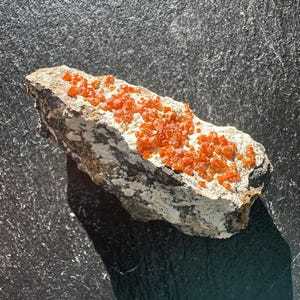 May include: A piece of rock with a light grey base, covered in small, bright orange crystals. The crystals are clustered together, creating a textured surface. The rock has a natural, earthy appearance, with variations in colour and texture.