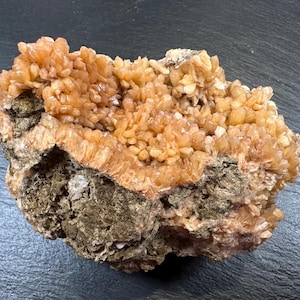 May include: A natural mineral specimen featuring a cluster of small, light orange crystals atop a darker, textured base. The crystals have a clustered, granular appearance, and the base has a rough, earthy texture.