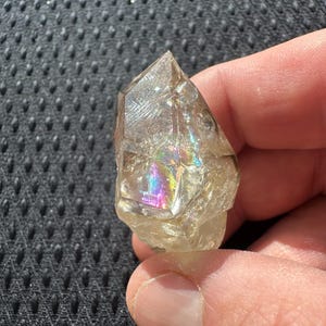 May include: A clear quartz crystal with a pointed tip and a rough, textured surface. The crystal displays a rainbow of colors where light refracts through it. The crystal is held in a hand.