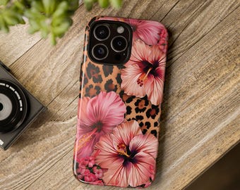 May include: A phone case featuring a vibrant floral design with pink hibiscus flowers and a leopard print background. The case has a glossy finish and a black camera lens cutout. The design is set against a wooden surface.