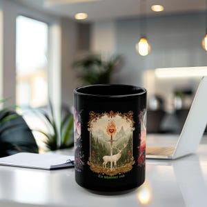 Puede incluir: Taza de cerámica negra con una ilustración detallada de un ciervo, una espada y un corazón con el texto "To whatever end". El diseño está enmarcado por bordes ornamentados y sobre un fondo montañoso. Un portátil y un bloc de notas están en el fondo.