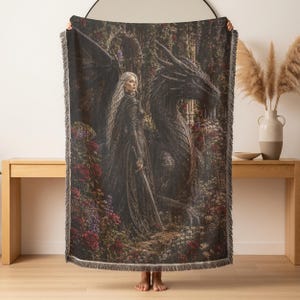 May include: A rectangular woven throw blanket featuring a detailed illustration of a long-haired figure in dark armor holding a sword, standing beside a large black dragon. The scene is set in a garden with red and white flowers and stone arches.