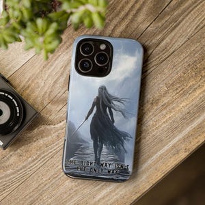 Puede incluir: Funda de teléfono con una ilustración dramática de una figura con pelo largo sosteniendo una espada, sobre un fondo montañoso. La funda tiene el texto "THE RIGHT WAY ISN'T THE ONLY WAY" en la parte inferior. La funda está sobre una superficie de madera.