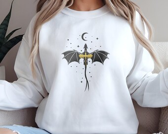 Puede incluir: Sudadera blanca con una silueta de dragón negro y dorado volando sobre un fondo de luna creciente y estrellas.