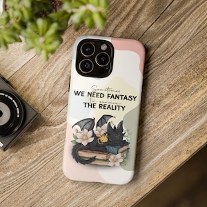 Könnte beinhalten: Handyhülle mit einem skurrilen Design mit einem schwarzen Drachen und einem gelben Drachen, die auf Büchern ruhen, umgeben von weißen Blumen. Die Hülle hat einen pastellfarbenen Hintergrund und den Text "Sometimes we need fantasy to survive the reality."