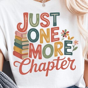 Puede incluir: Camiseta blanca con el texto "JUST ONE MORE CHAPTER" en letras coloridas. El diseño incluye una pila de libros y elementos florales. Un estilo casual.