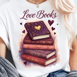 May include: White t-shirt with the text "Love Books" in burgundy script. The shirt features a stack of illustrated books in shades of purple and red, with a heart design on the top book. Small heart accents are scattered around the books.