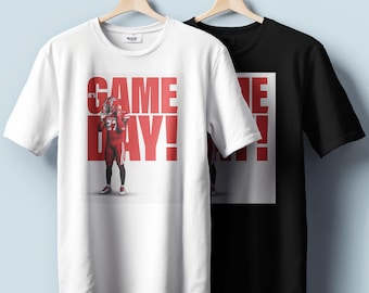 May include: Two t-shirts hanging on wooden hangers. One is white, the other is black. The white t-shirt has a large red graphic that says "IT'S GAME DAY!" with a football player in the center. The black t-shirt has a partial graphic.
