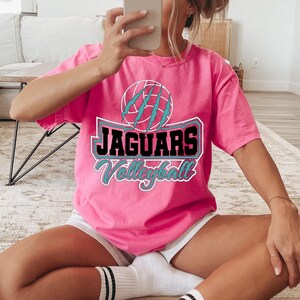 May include: A pink t-shirt with the text "JAGUARS Volleyball" in a teal and white design. The shirt features a graphic of a volleyball with claw marks. The shirt is short-sleeved.