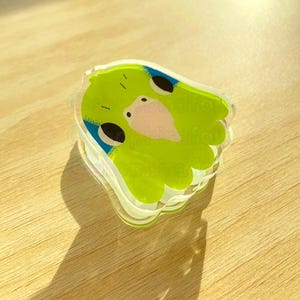 May include: A green and blue resin charm featuring a cartoon parakeet with a pink beak and black eyes. The charm is shaped like a parakeet and has a clear resin finish.