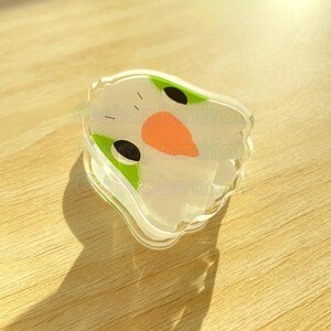 May include: A clear plastic charm with a white bird face, green eyes, and an orange beak. The charm is shaped like a bird and has a white background.