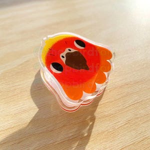 May include: A clear acrylic keychain with a red and yellow bird design. The bird has a brown beak and black eyes.