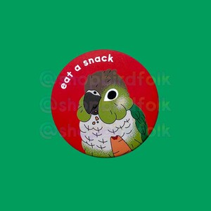 May include: A red circle button with the text "eat a snack" in white. A green and brown Quaker parrot is depicted on the button, holding a carrot in its beak.