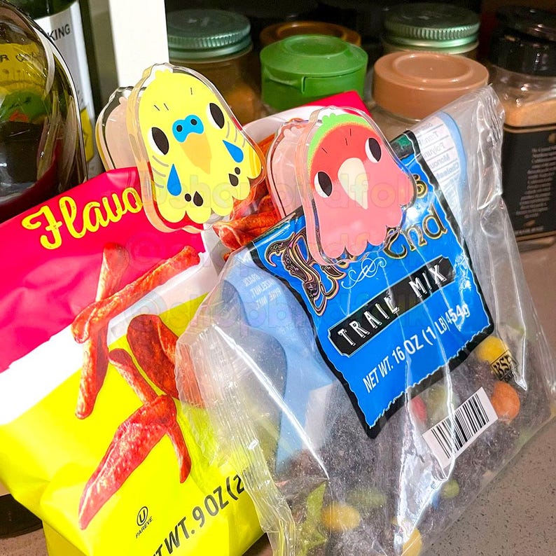 May include: Two colorful plastic bag clips shaped like cartoon birds. The yellow bird clip is on a bag of red and yellow flavored tortilla chips. The pink bird clip is on a bag of trail mix. The bag of trail mix has the text "Trail Mix" and "Net Wt. 16 oz (1 lb 454g)".