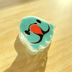 May include: A light blue resin charm with a cartoon bird design. The bird has a red beak and black eyes. The charm is shaped like a bird with its wings spread out.
