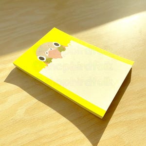 Puede incluir: Un bloc de notas amarillo con un lindo pájaro de dibujos animados asomándose por la parte superior. El bloc de notas tiene un área blanca en blanco para escribir.