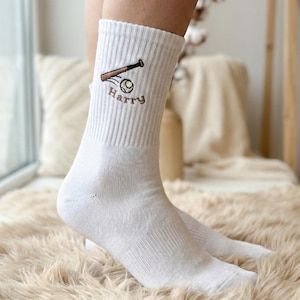 Op de afbeelding: Witte sokken met een geborduurd ontwerp van een honkbalknuppel en -bal. Het woord "Harry" is onder de honkbalafbeelding geborduurd. De sokken zijn gemaakt van een zacht, geribbeld materiaal.