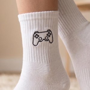 May include: White crew socks with a black embroidered video game controller design. The socks are ribbed and appear to be made of a soft, comfortable material. The socks are on a fluffy, white rug.