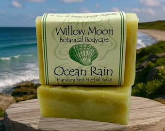 May include: Two bars of handcrafted herbal soap, one stacked on top of the other, with a label that reads "Willow Moon Botanical Bodycare Ocean Rain." The label features a green seashell graphic. The soap is a light yellow color and rests on a wooden surface.