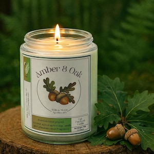 May include: A lit glass jar candle with a label that reads "Amber & Oak" and features an acorn illustration. The candle is on a wooden surface with oak leaves and acorns. The candle is 7.5 ounces.