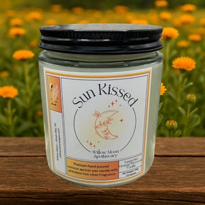 May include: A glass jar candle with a black lid, labeled "Sun Kissed" with a crescent moon design. The label also reads "Willow Moon Apothecary" and lists the scent as Tangerine Orange Blossom Vanilla. The candle weighs 7.8 ounces.
