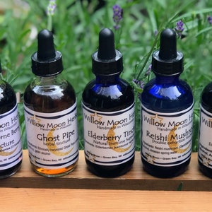 May include: Seven glass bottles with dropper tops, each containing a different colored liquid. The bottles are labeled with the names of various herbs and plants, including Hawthorn Berry, Ghost Pipe, Elderberry, Reishi Mushroom, Chaga, and St. Johns Wort. The bottles are arranged in a row on a wooden surface.