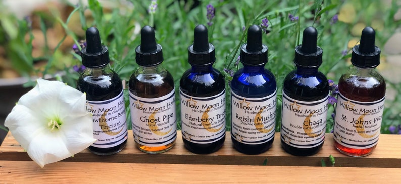 May include: Seven glass bottles with dropper tops, each containing a different colored liquid. The bottles are labeled with the names of various herbs and plants, including Hawthorn Berry, Ghost Pipe, Elderberry, Reishi Mushroom, Chaga, and St. Johns Wort. The bottles are arranged in a row on a wooden surface.