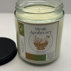 May include: A clear glass jar candle filled with off-white wax, featuring a label with the text Mystic Apothecary and an illustration of a mortar and pestle. The label lists Peppercorn, Sage, Balsam, Chamomile and a net weight of 7 ounces.