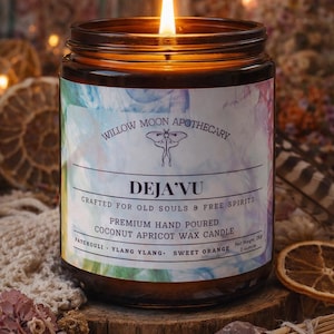 May include: A lit candle in a glass jar with a label that reads "DEJA'VU" from Willow Moon Apothecary. The label lists ingredients and the candle is made with coconut apricot wax. The candle is on a wooden surface.
