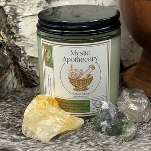 Willow Moon Apothecary’s Mystic Apothecary candle in a clear glass jar with black lid and earthy green label. The front features a golden illustration of herbs, mortar and pestle, and stars. Surrounded by raw crystals and a wooden mortar, the candle rests on faux fur with birch bark in the background. Scent notes include peppercorn, sage, balsam, and patchouli, creating a grounding and magical aroma.