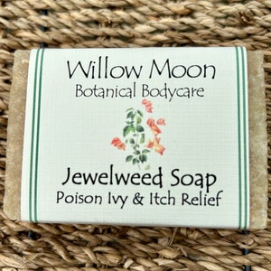 以下が含まれることがあります： 「Willow Moon Botanical Bodycare Jewelweed Soap Poison Ivy & Itch Relief」と書いてある白いラベルの石鹸。ラベルは緑色の縁取りと花のイラストが描かれている。