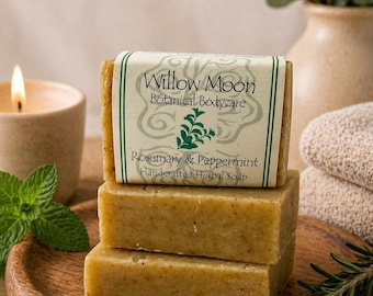 May include: Three bars of handcrafted herbal soap, labelled "Willow Moon Botanical Bodycare: Rosemary & Peppermint." The soaps are stacked on a wooden plate, with a lit candle and sprigs of mint and rosemary. The soap bars are approximately 10 cm long.
