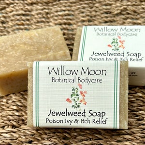 Three bars of Jewelweed Soap from Willow Moon Botanical Bodycare displayed on a woven natural mat. The front bar features a clean label with green stripes and a watercolor jewelweed illustration, labeled for poison ivy and itch relief. Bars have a rustic, handcrafted texture in a soft amber tone.