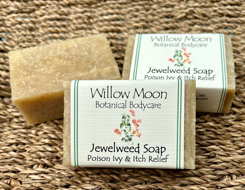 Three bars of Jewelweed Soap from Willow Moon Botanical Bodycare displayed on a woven natural mat. The front bar features a clean label with green stripes and a watercolor jewelweed illustration, labeled for poison ivy and itch relief. Bars have a rustic, handcrafted texture in a soft amber tone.
