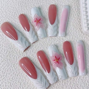 May include: Ten long, almond-shaped artificial nails with a white base and a pink, sculpted design. Each nail features a delicate, white, 3D floral design and a pink flower accent.