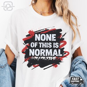 May include: White t-shirt with the slogan "NONE OF THIS IS NORMAL" in bold white text, set against a black and red abstract design. The shirt has a crew neck and short sleeves. The design includes a silhouette of a crowd at the bottom.