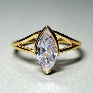 Könnte beinhalten: Ein goldener Ring mit einem klaren Stein in Marquise-Form, der in der Mitte eingesetzt ist. Der Ring hat ein geteiltes Schienen-Design.