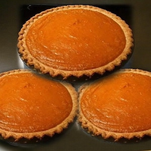 May include: Three pumpkin pies with golden-brown crusts and a smooth, orange filling. The pies are arranged on a dark surface, showcasing their round shape and crimped edges. A classic dessert.