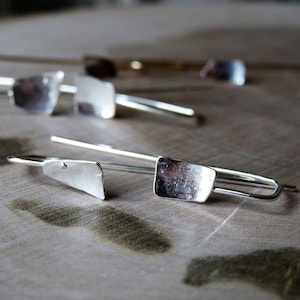 May include: Silver earrings with a rectangular shape and a slightly curved design. The earrings are on a wooden surface.