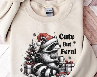 May include: A sand-coloured sweatshirt featuring a raccoon in a Santa hat, holding a mug, with gifts and a Christmas tree. The text reads "Cute But Feral." The sweatshirt has long sleeves and the text "Trendy Custom Sweaters Made in Texas."