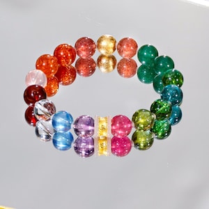 May include: A colorful beaded bracelet with a variety of round beads in shades of red, orange, gold, green, blue, and purple. The beads are strung together with a gold-colored spacer. The bracelet is displayed on a reflective surface.