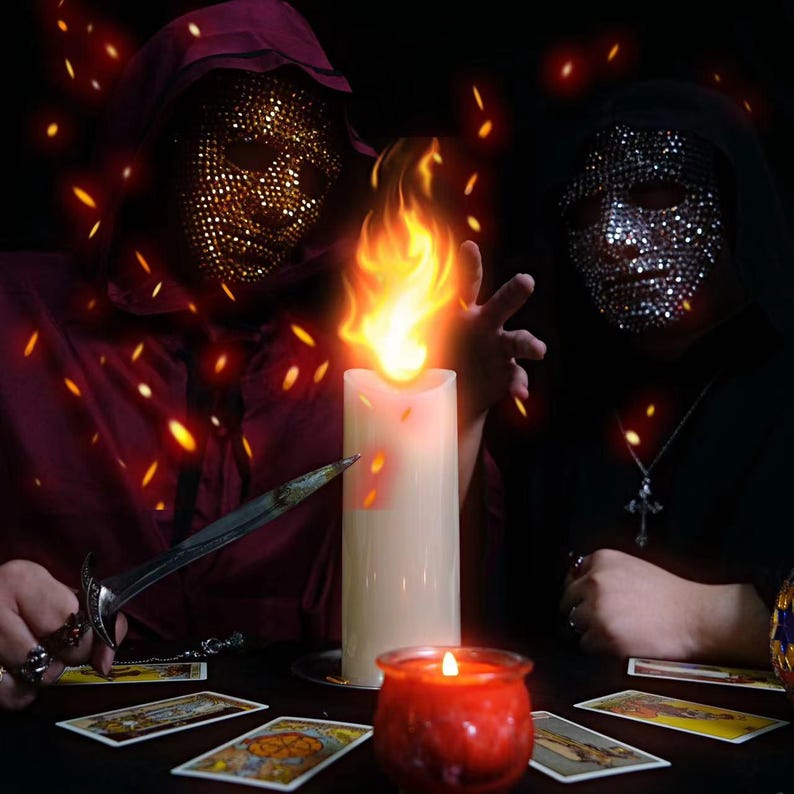 K&ouml;nnte beinhalten: Zwei Figuren in Kapuzenroben und geschm&uuml;ckten Masken sind um eine gro&szlig;e Kerze mit einer Flamme positioniert. Ein Dolch und Tarotkarten liegen auf dem Tisch. Eine kleine rote Kerze brennt in der N&auml;he, mit Funken und einem dunklen Hintergrund.