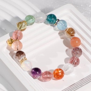 Może przedstawiać: Kolorowa bransoletka z różnorodnymi koralikami z kamieni szlachetnych w odcieniach zieleni, różu, pomarańczy i fioletu. Bransoletka zawiera złote akcenty i jest prezentowana na białej powierzchni.