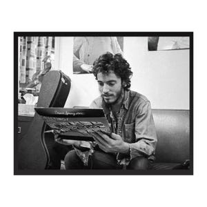 May include: Black and white photograph of a man seated, holding and looking at a record album. The album cover is visible, with text that reads "Bruce Springsteen" and "Greetings". A guitar case is visible in the background.