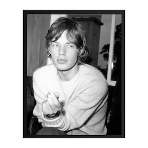 May include: Black and white portrait of a person with long hair wearing a light-colored sweater. The person is looking towards the camera, with a slight smile. The image is framed in a black border.